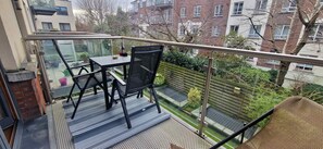 A balcony with a table and chairs, a glass railing, and a view of a garden and buildings.