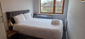 A neatly made bed with white linens, a bedside lamp, and a window with a view of buildings outside.