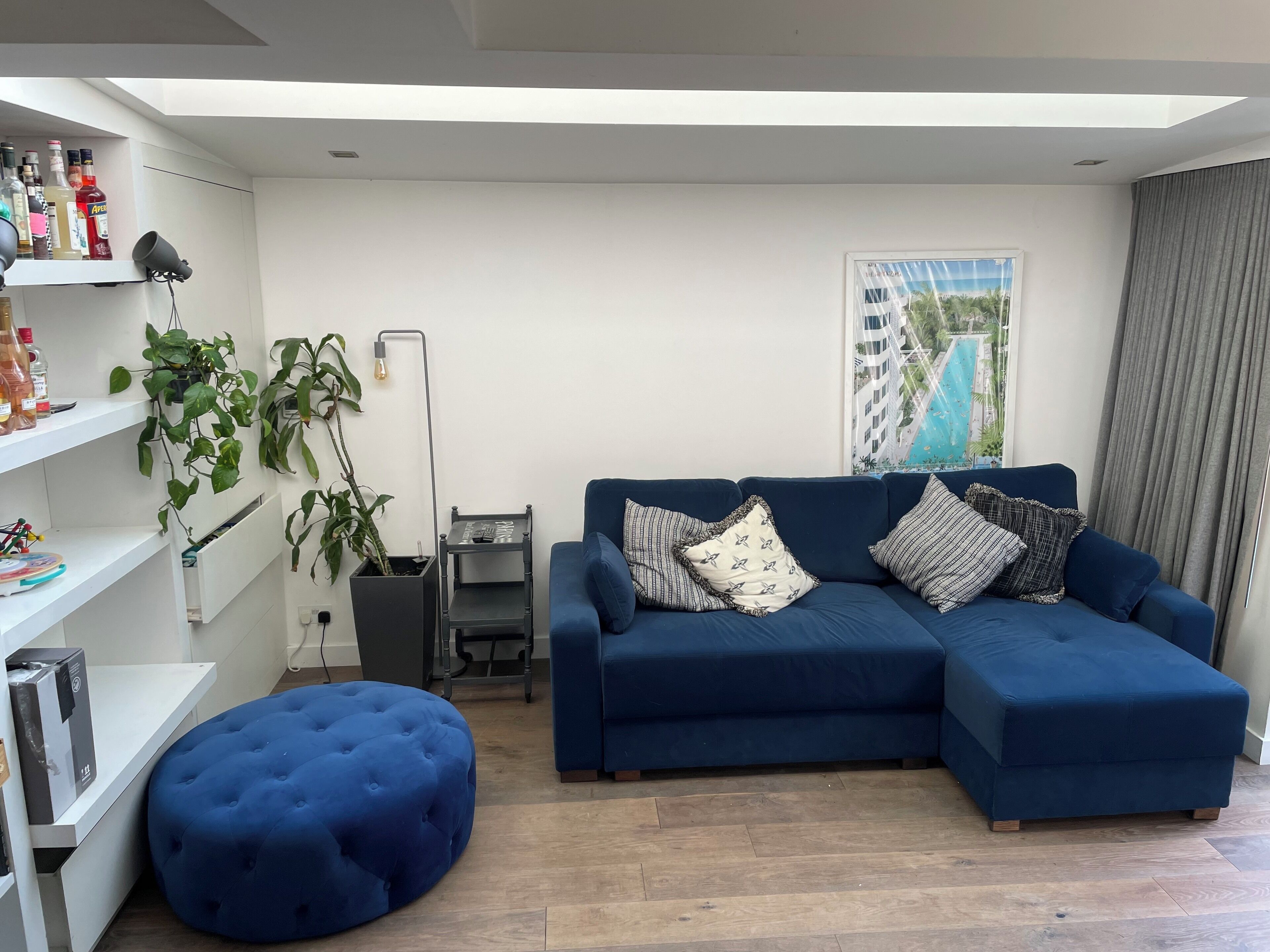 A modern living room with a blue sectional sofa, a round ottoman, a white shelving unit with various items, a floor lamp, and a large window with a view of buildings.