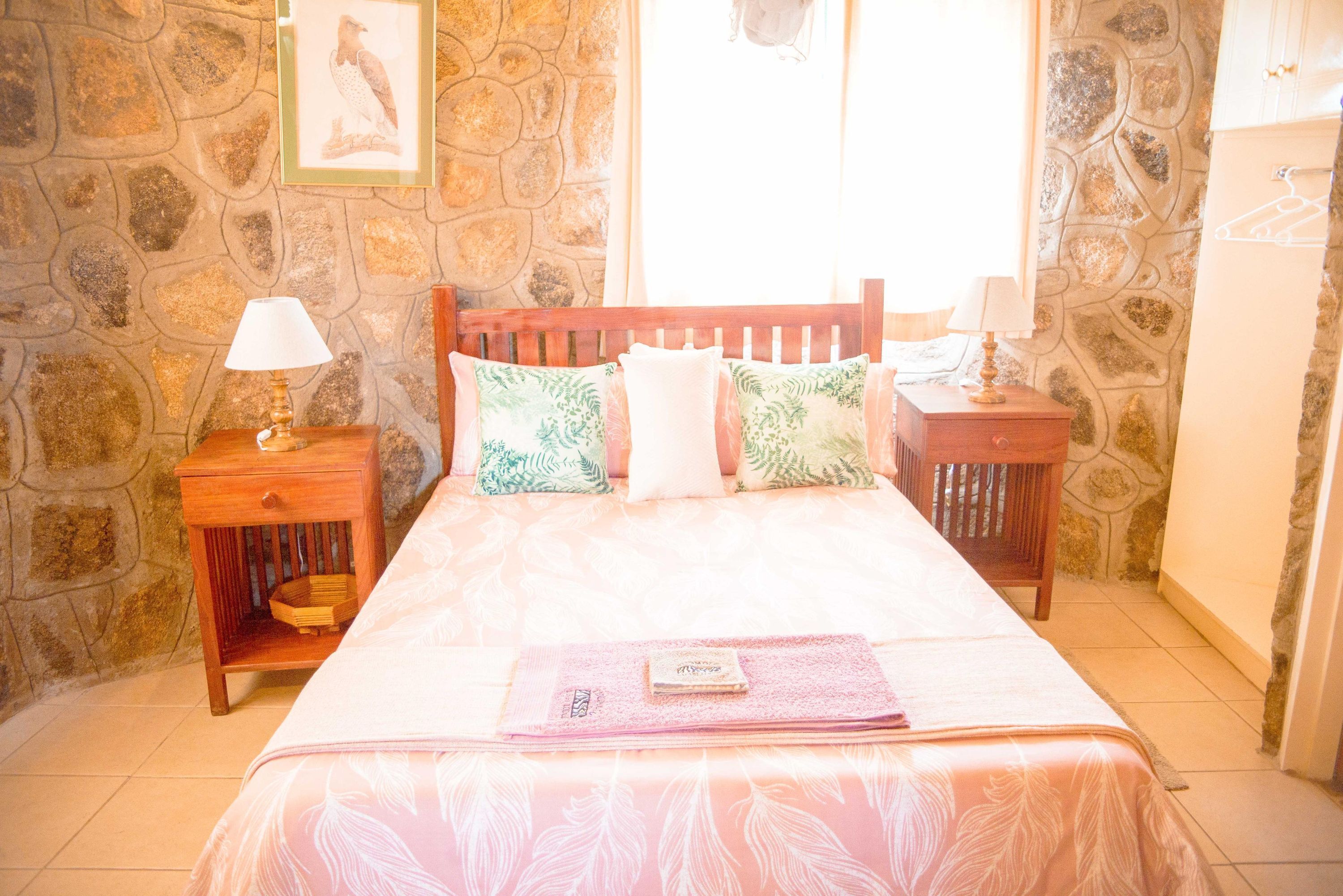 Une chambre avec un mur en pierre, un cadre de lit en bois, deux tables de chevet avec des lampes et une fenêtre avec des rideaux.