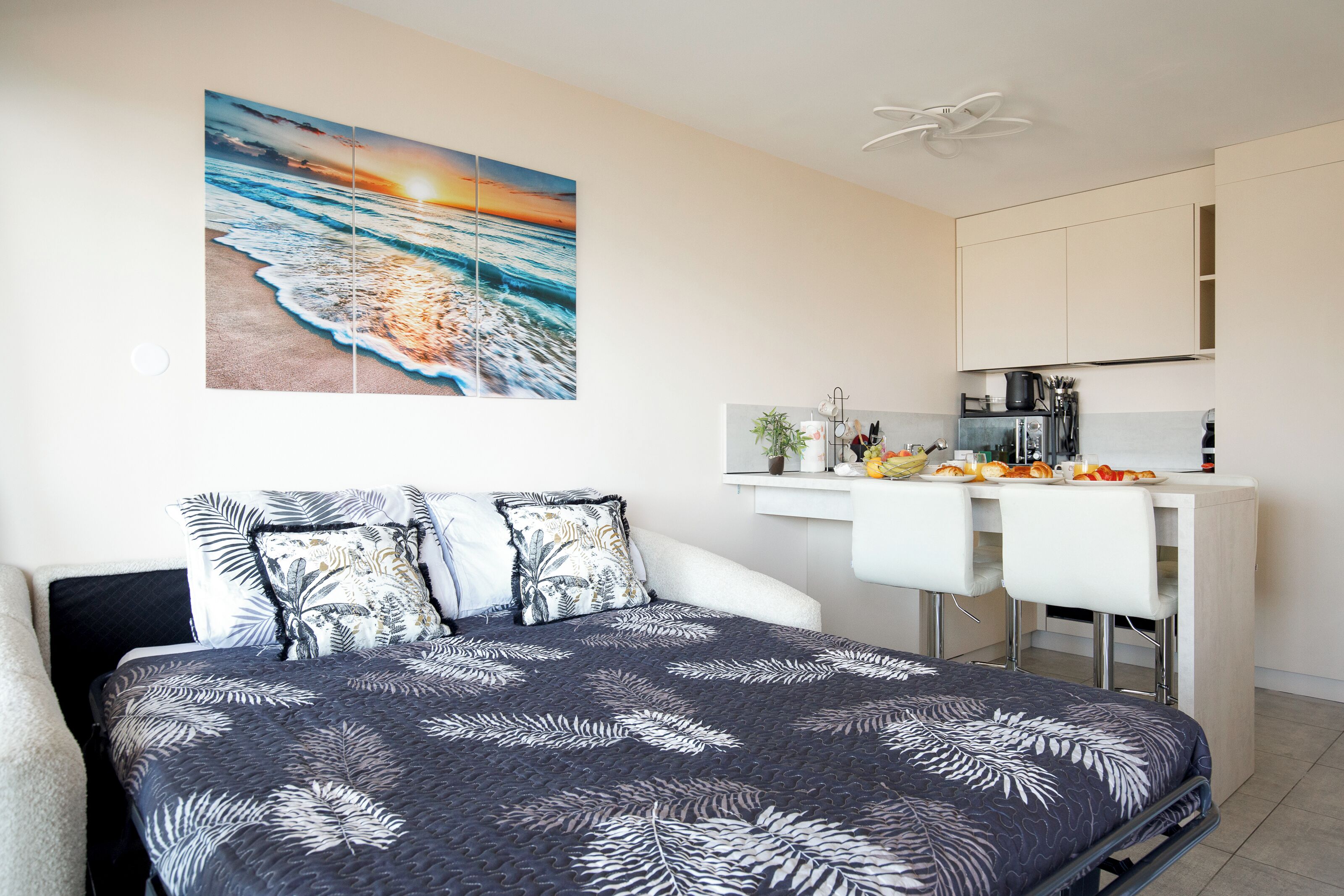 Une chambre moderne avec un lit, un tableau représentant un coucher de soleil sur l’océan, une kitchenette équipée d’une cuisinière et un ventilateur de plafond blanc.