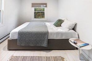A modern bedroom with a bed, bedside table, and a window with blinds. - Charming 2-bedroom cottage in Sag Harbor with AC, WiFi (Sag Harbor)