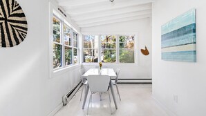 A bright dining area with a white table and chairs, large windows with a view of trees, and a wall with a geometric art piece. - Charming 2-bedroom cottage in Sag Harbor with AC, WiFi (Sag Harbor)