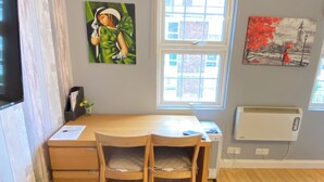 Une pièce comprenant un bureau en bois, une chaise, un radiateur et deux fenêtres équipées de stores.