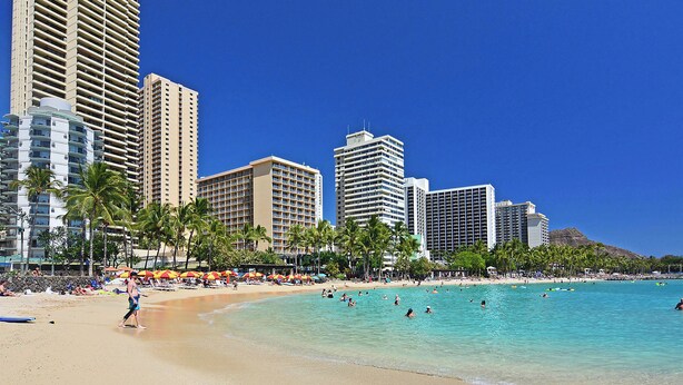 A beach with clear turquoise waters, palm trees, and tall buildings in the background.