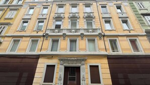 Um edifício com vários andares e detalhes arquitetônicos ornamentados, incluindo molduras decorativas e estátuas sobre as janelas.
