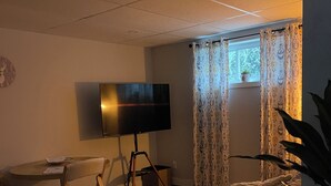 Un salon avec un téléviseur à écran plat posé sur un meuble, un canapé, une table à manger avec des chaises et une fenêtre agrémentée de rideaux à motifs.