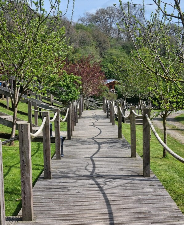 Una pasarela de madera con barreras de cuerda, rodeada de árboles y césped verdes.