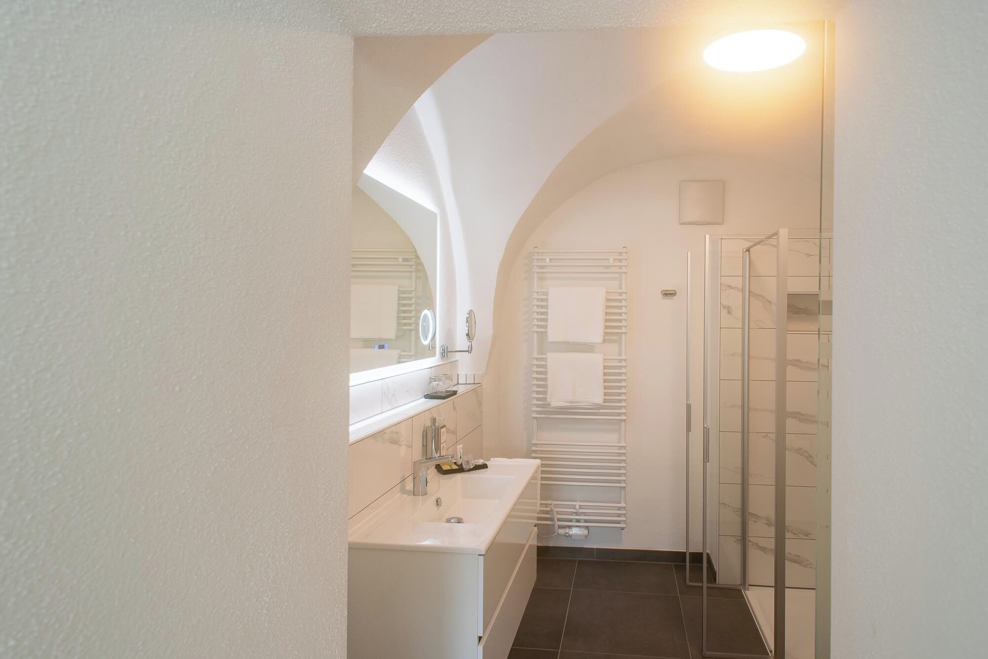 Ein schmaler Flur führt zu einem Badezimmer mit Waschbecken, Spiegel und Dusche.