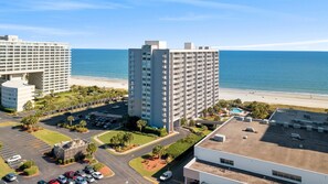 A high-rise hotel with a pool and beach view, parking lots, and a landscaped area.