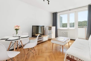 A modern hotel room with a wooden floor, a bed with a white duvet, a TV on a wooden stand, a white desk with a chair, and a large window with curtains.