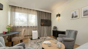 Un salon moderne avec un téléviseur à écran plat, une table à manger avec des chaises, un canapé, une table basse avec des livres et des tasses, et une grande fenêtre avec des rideaux.