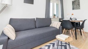 Un salon moderne avec un canapé gris, une table à manger avec des chaises, une lampe et une fenêtre avec des rideaux.