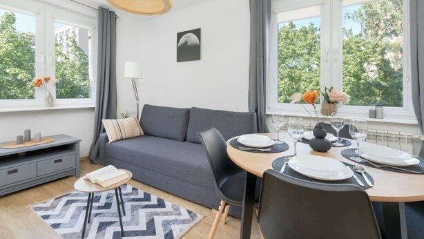 Un salon moderne avec un canapé gris, une table à manger dressée pour deux personnes et une grande fenêtre donnant sur un paysage arboré.