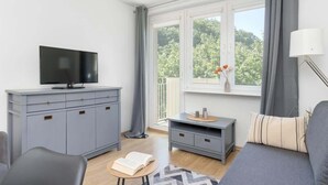 Un salon moderne avec un canapé gris, un téléviseur posé sur un meuble, une petite table avec un livre, et une vue sur la verdure par la fenêtre.