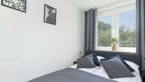Une chambre à coucher avec un lit, une fenêtre avec des rideaux, un tableau accroché au mur et une petite plante posée sur le rebord de la fenêtre.