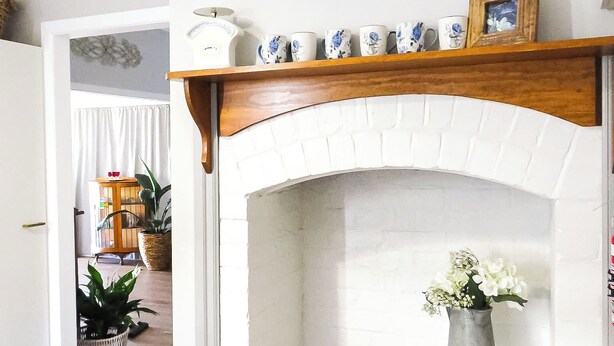 Una habitación con chimenea de ladrillo blanco, repisa de madera con objetos decorativos y un cuadro colgado encima.