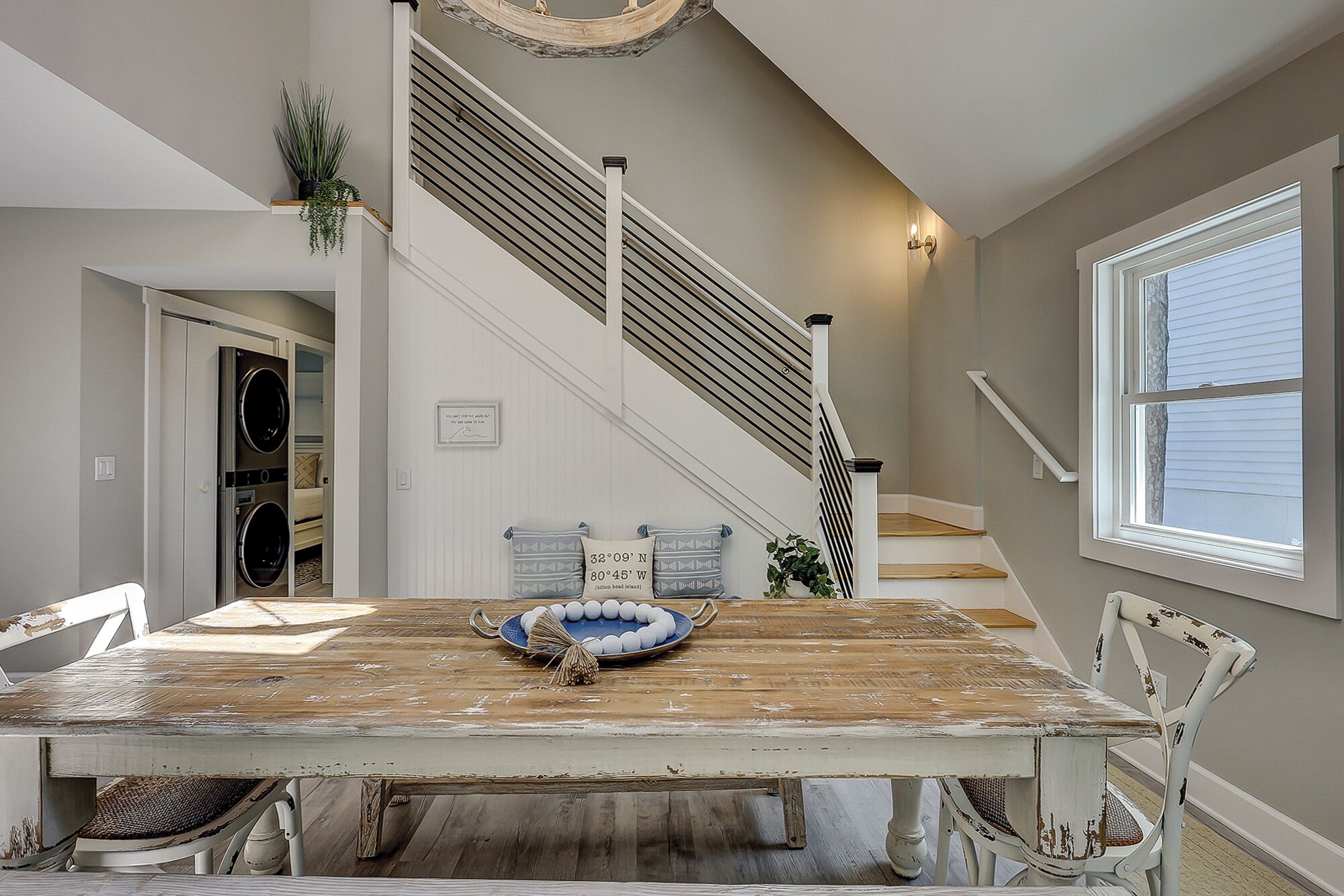 A dining area with a rustic wooden table, white chairs, a blue and white decorative plate, and a staircase with a striped railing.