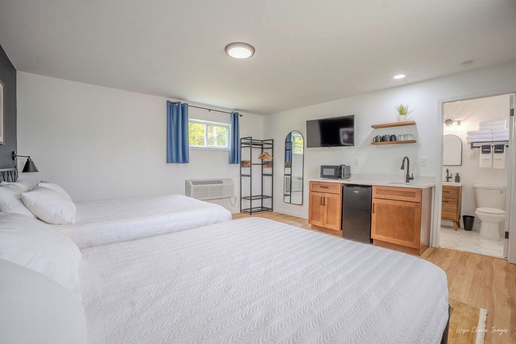 A modern hotel room with two beds, a kitchenette, a TV, and a bathroom.