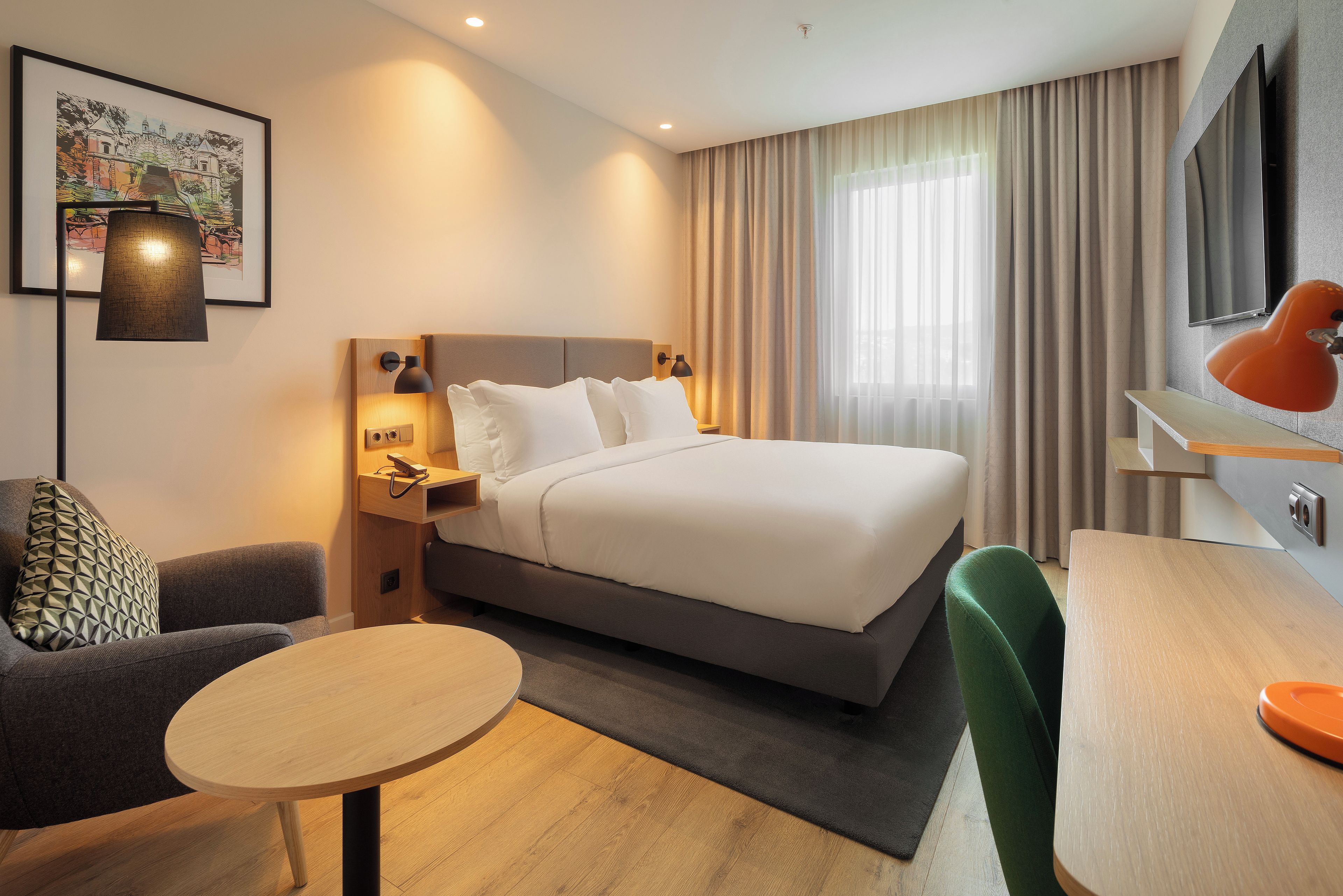 A modern hotel room with a large bed, a desk, a chair, a small table, and a TV.