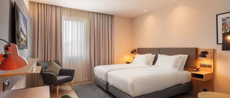 A hotel room with a bed, bedside tables, a desk, a chair, and a window with curtains.