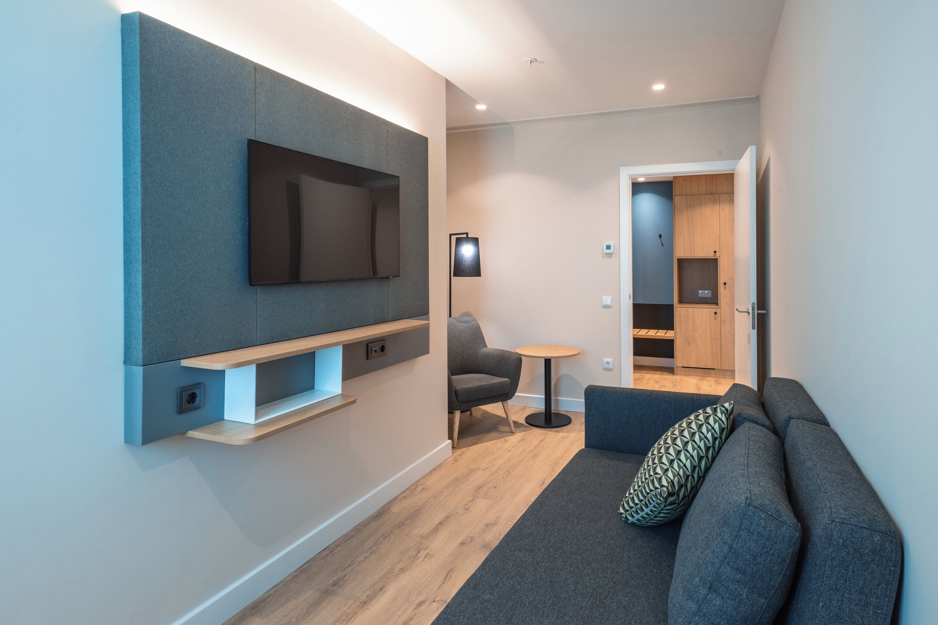 A modern living room with a flat-screen TV mounted on a wall, a grey sofa with a patterned pillow, a small round table, and a wooden cabinet in the background.