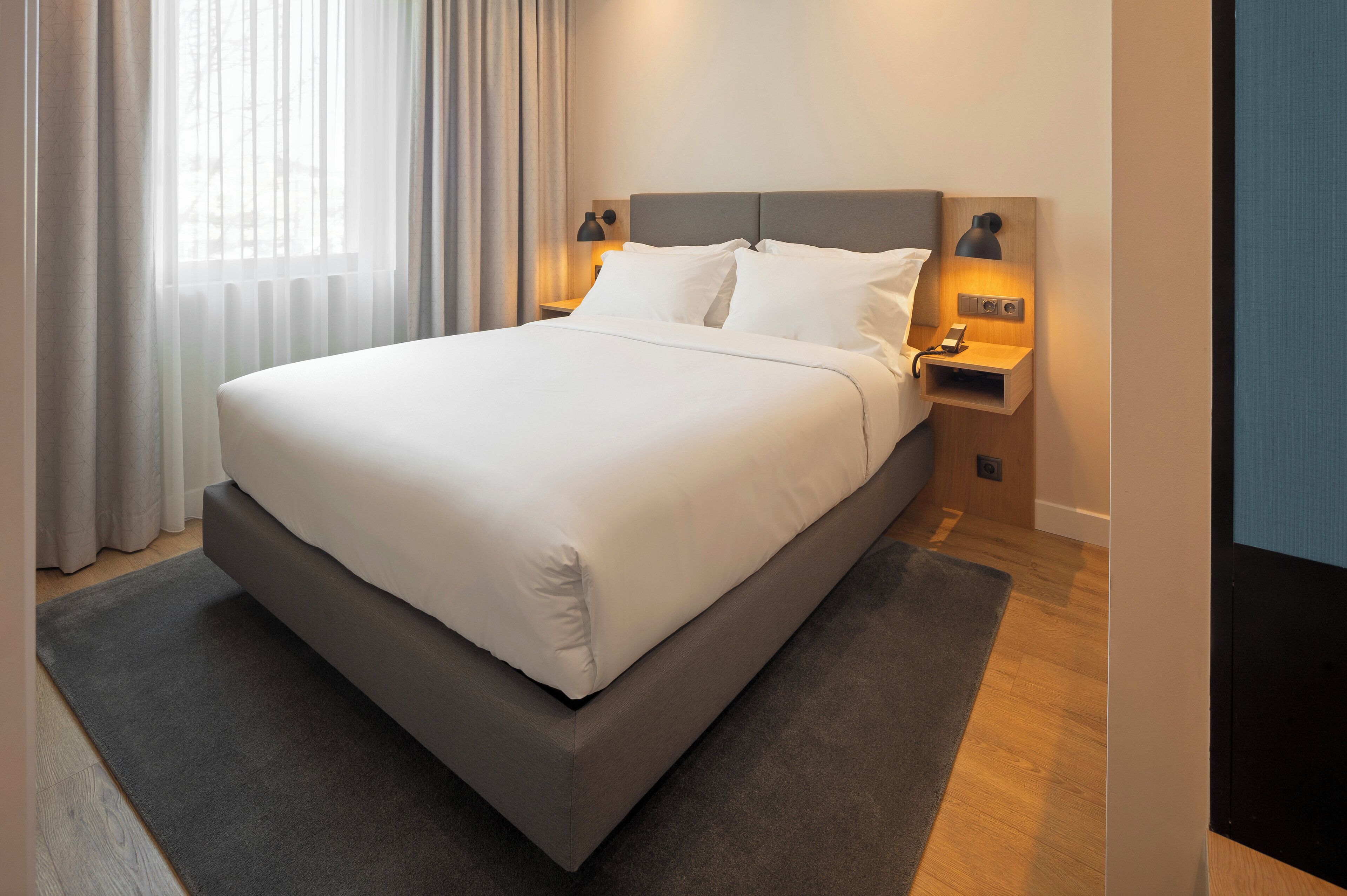 Ein modernes Hotelzimmer mit einem großen Bett, zwei Nachttischen mit Lampen, einem grauen Kopfteil und Blick auf ein Gebäude durch leichte Vorhänge.