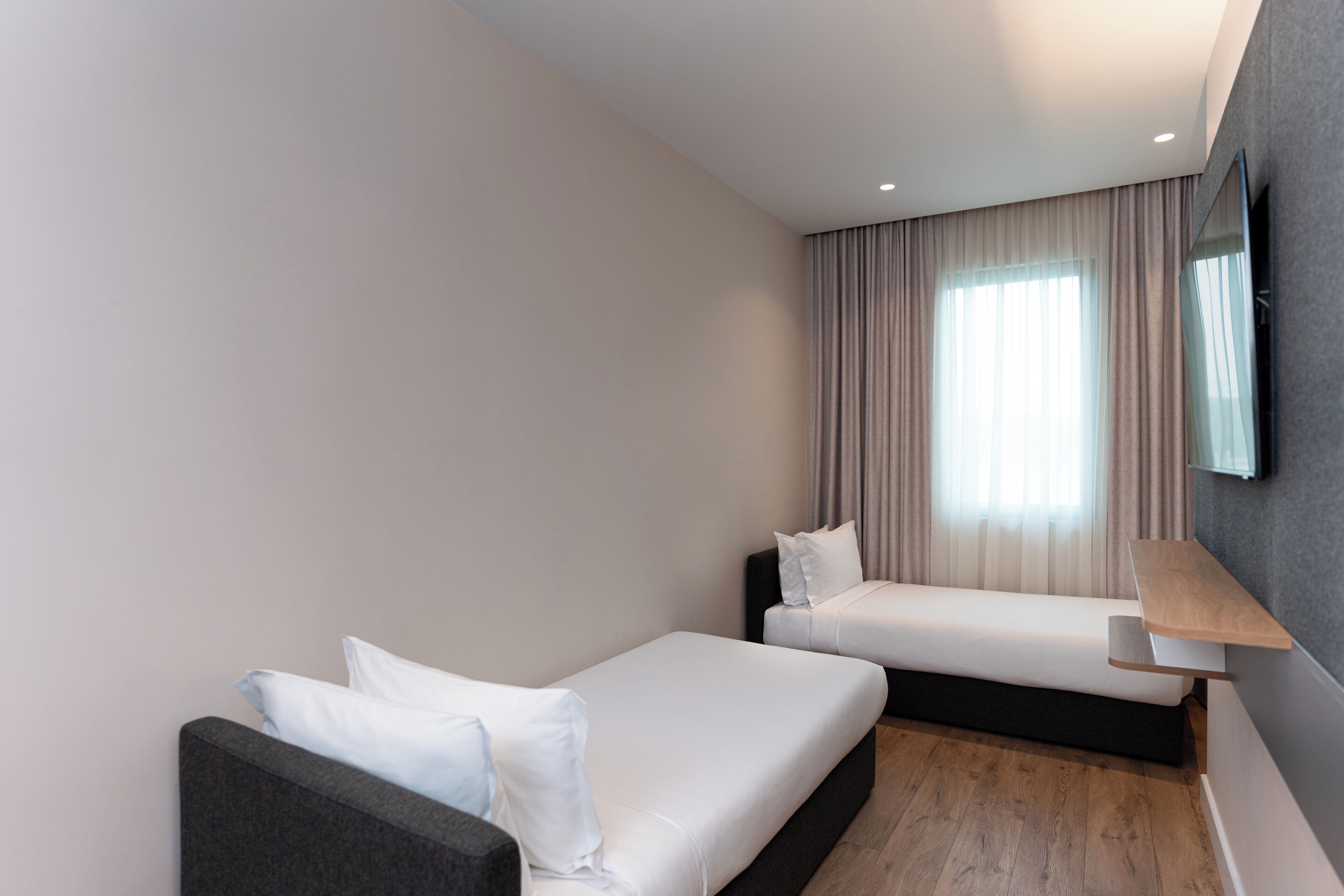 A modern hotel room with a sofa, a bed, a TV mounted on the wall, and a window with curtains.