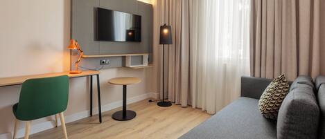 Una habitación de hotel moderna con televisión de pantalla plana, un escritorio con lámpara, una silla verde, un sofá con cojines estampados y un ventanal con cortinas.