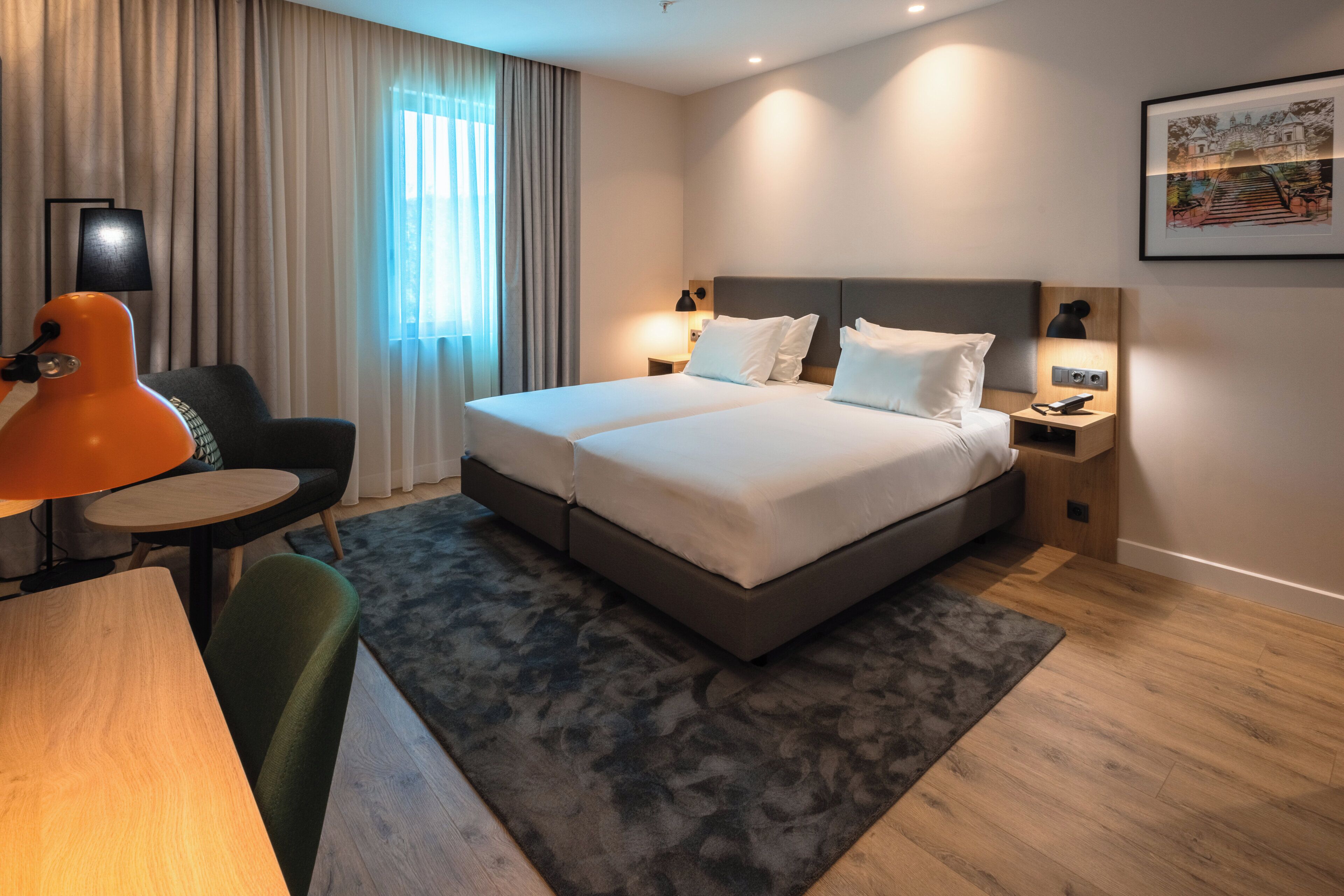 A hotel room with a double bed, a desk with a chair, a lamp, a bedside table with a phone, and a painting on the wall.