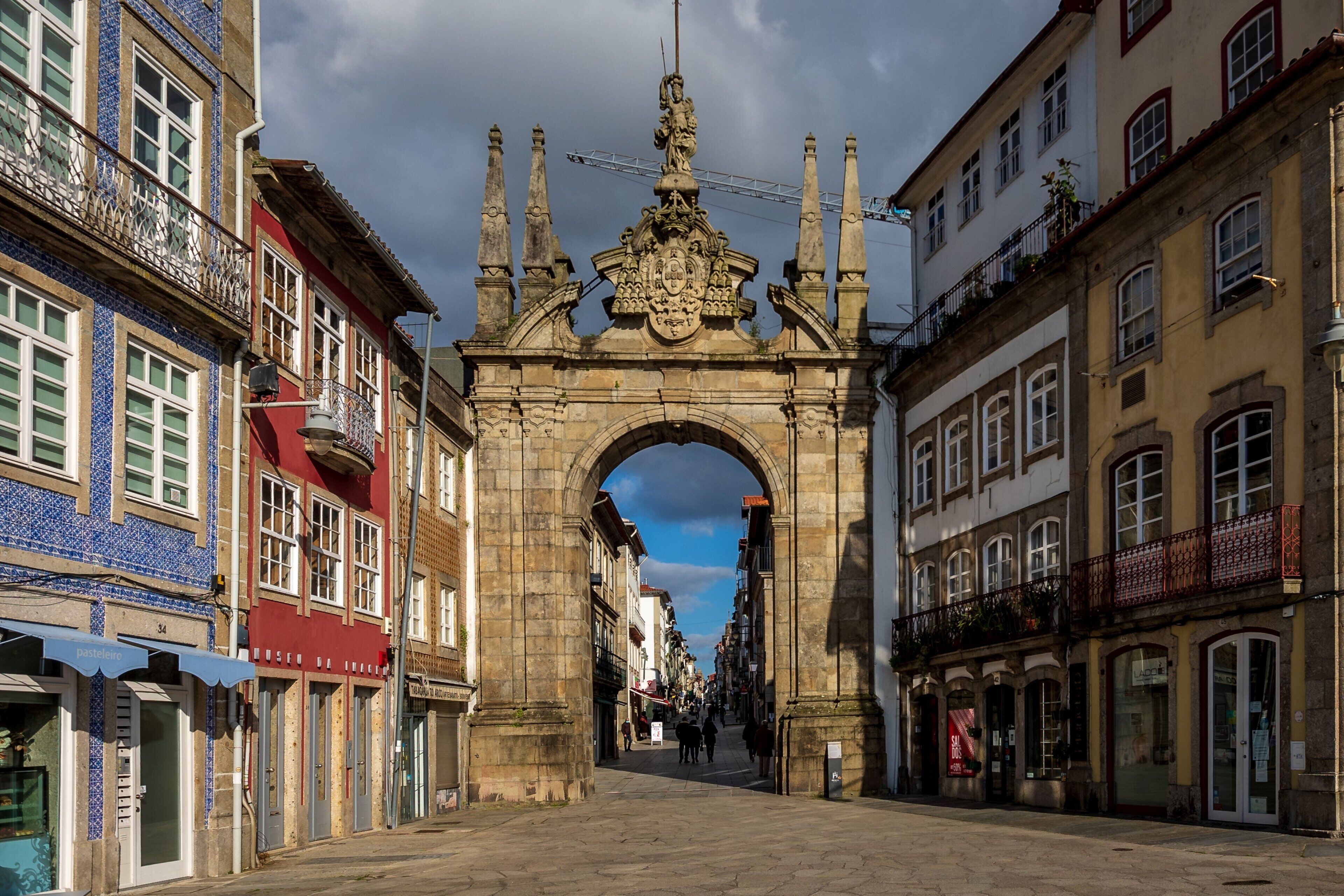 Ein historischer Steinbogen in einem europäischen Stadtplatz, umrahmt von farbenfrohen Gebäuden mit Balkonen und Schaufenstern.