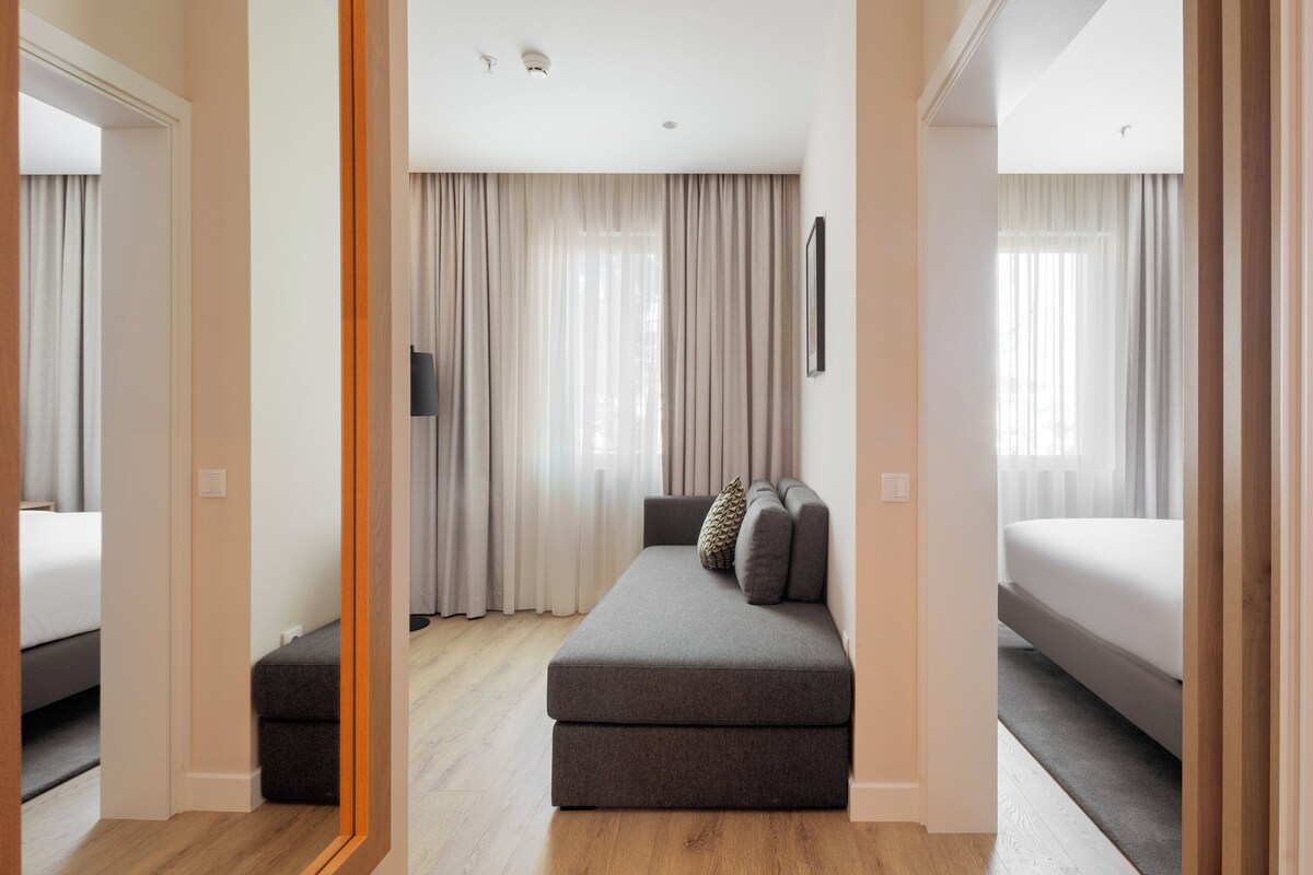 Una habitación de hotel moderna con un sofá gris, un televisor de pantalla plana y un ventanal con cortinas.