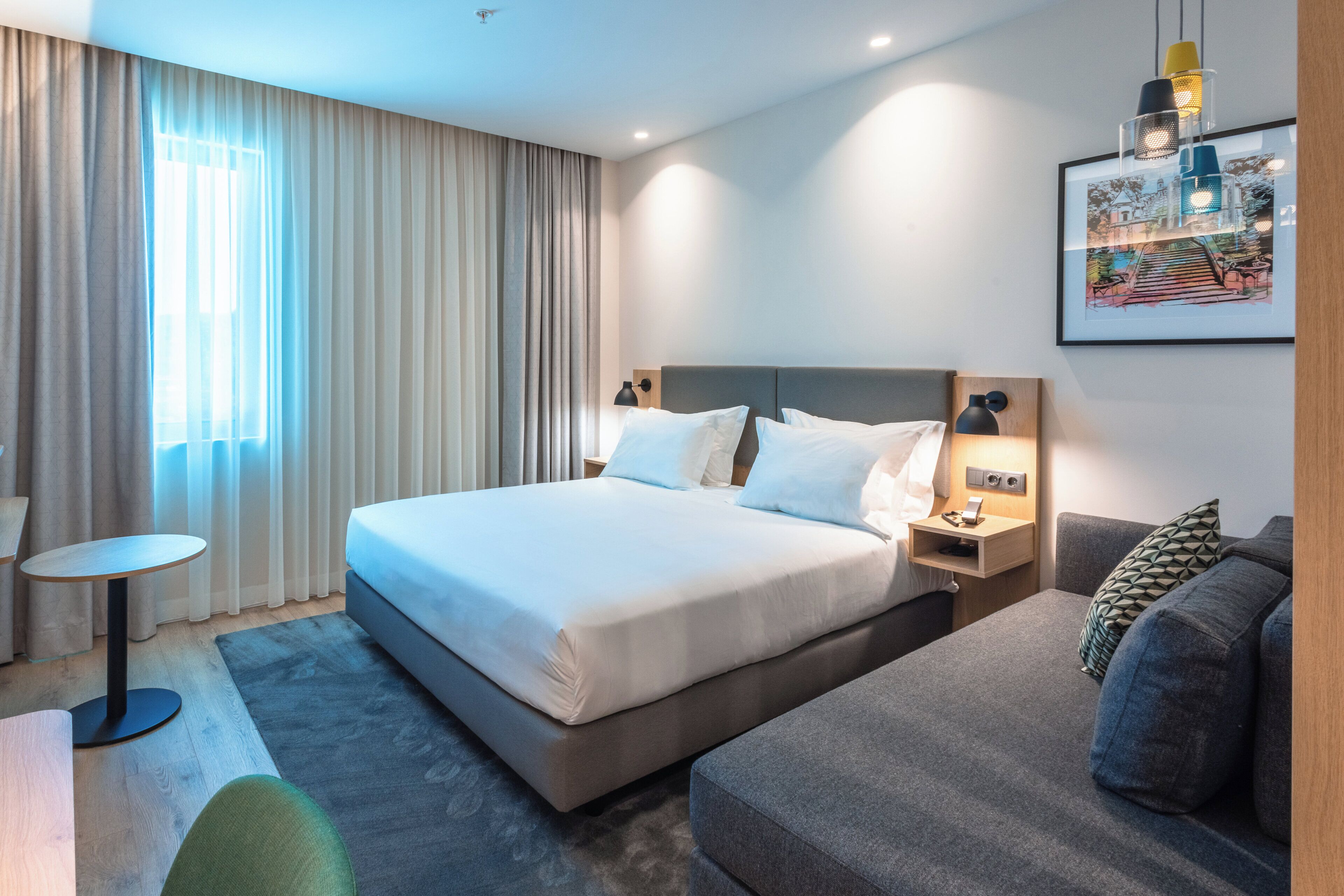 A hotel room with a large bed, a bedside table, a chair, a sofa, and a painting on the wall.