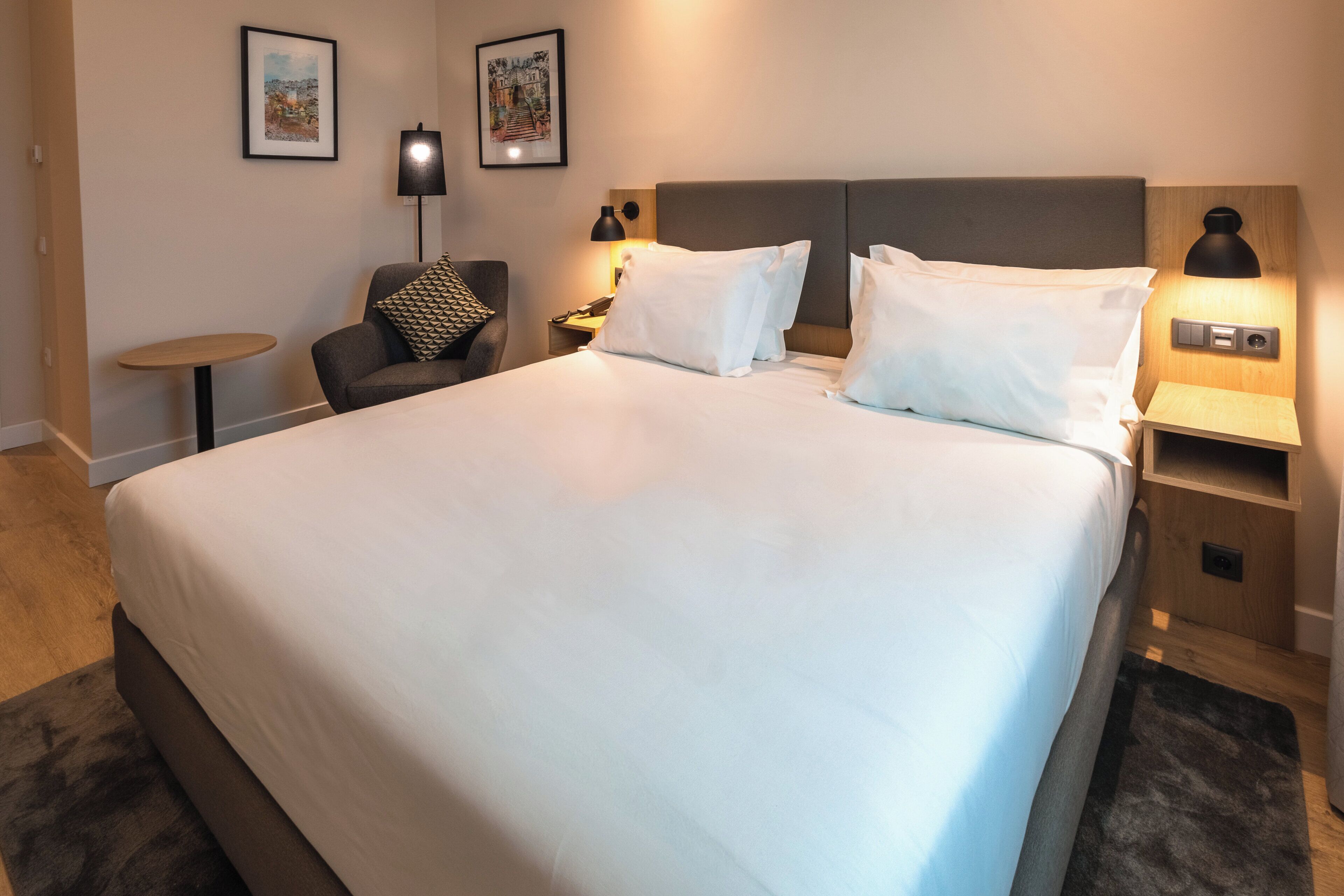 Et hotelværelse med en stor seng, en stol, et natbord og to væglamper.