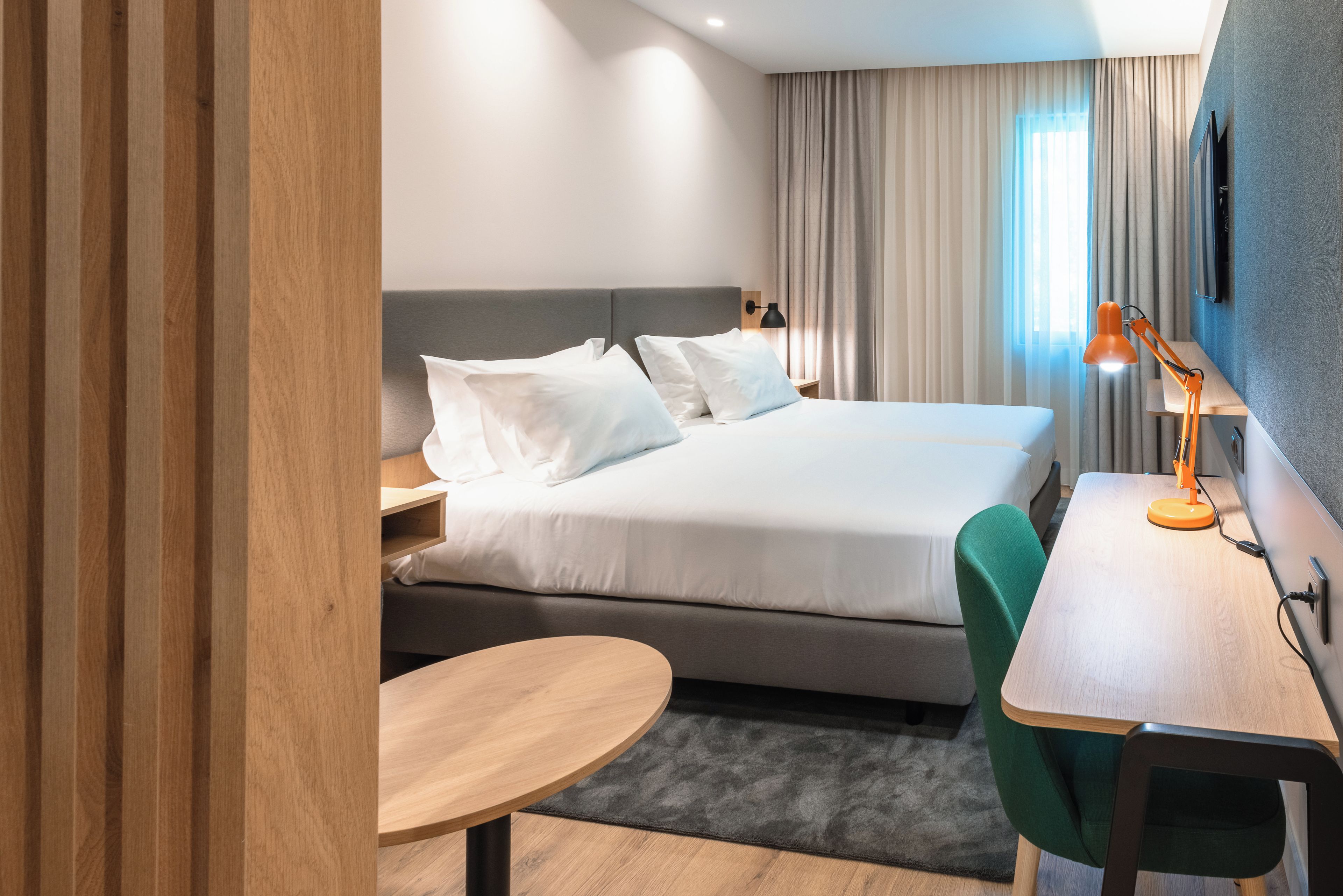 A hotel room with a bed, bedside table, lamp, and a desk with a chair.