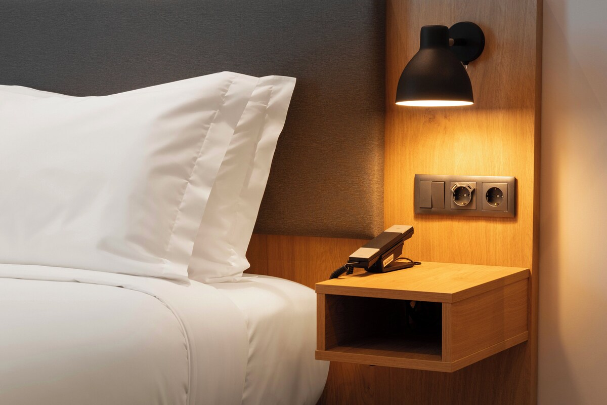 Una cama bien hecha con sábanas blancas, una mesita de noche con una lámpara y un teléfono en la pared.