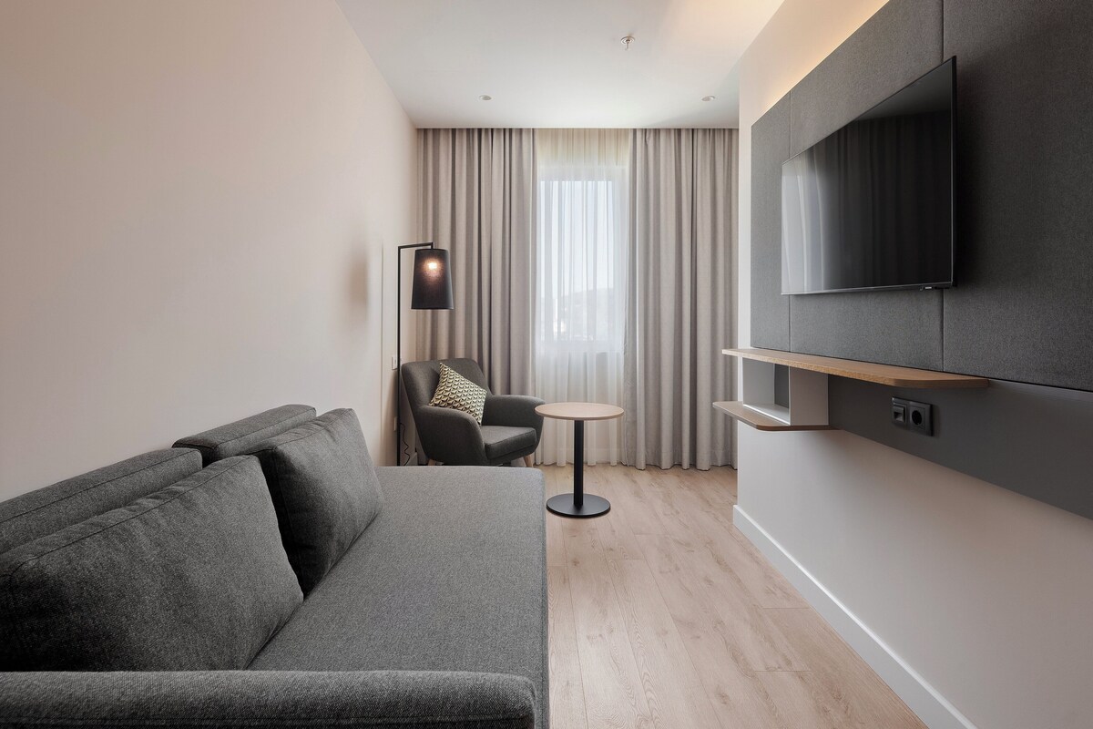 A modern hotel room with a grey sofa, a small round table, a chair, and a TV mounted on the wall.