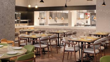 A modern restaurant with multiple dining tables and chairs, a kitchen area in the background, and pendant lighting.