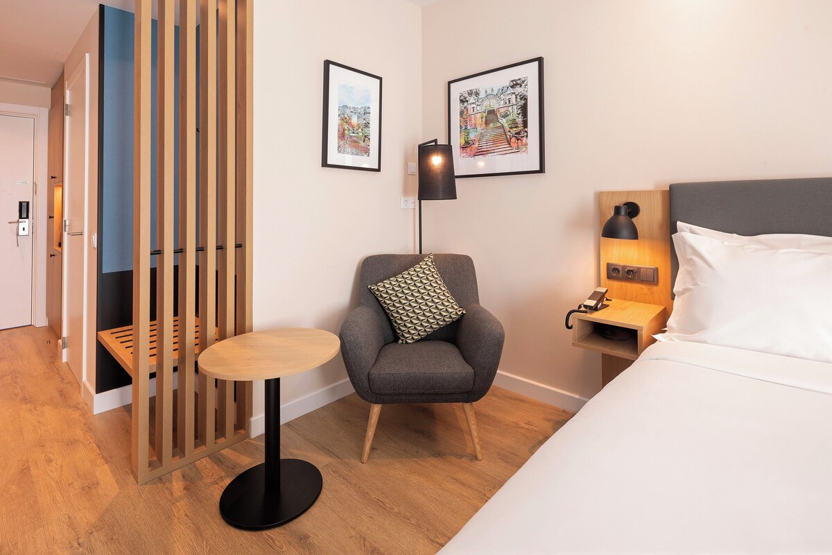 Hotelkamer met een bed, een stoel, een klein tafeltje, een nachtkastje en twee ingelijste foto's aan de muur.
