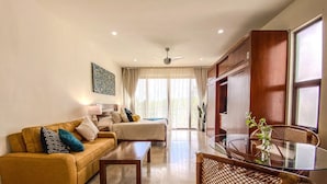 Una sala moderna con sofá, una mesa de centro y una mesa de comedor con cubierta de vidrio. Se ve un ventilador de techo, un ventanal con cortinas y un mueble de madera.