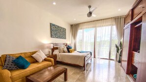 Habitación de hotel con cama, sofá, mesa de centro, ventilador de techo y un ventanal con cortinas.
