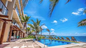 Zona de piscina en un resort con vista despejada al océano, palmeras y un edificio con balcones.