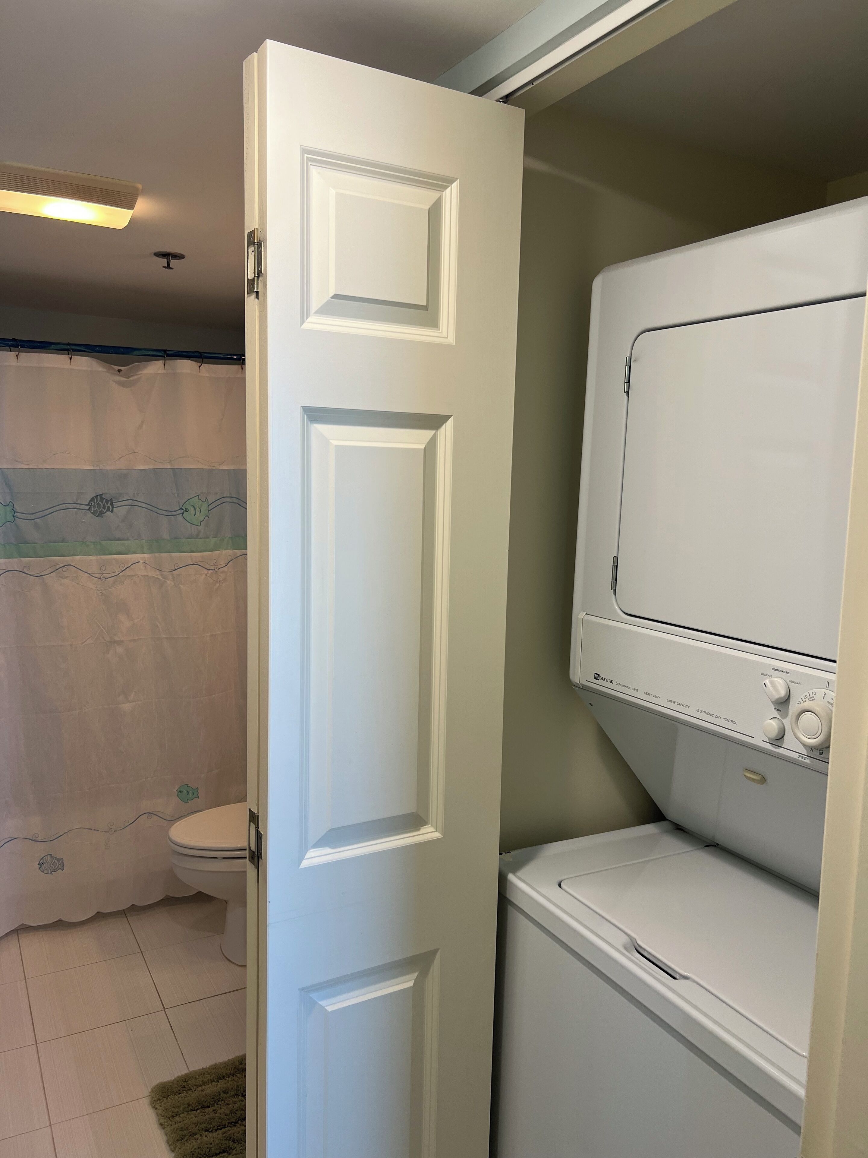 A bathroom with a white door, a toilet, a shower curtain, and a stacked washer and dryer.