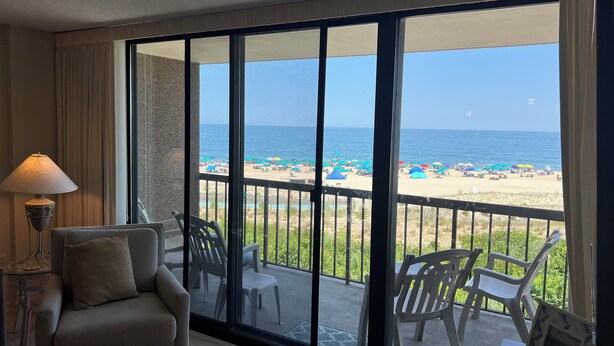 A balcony with a view of a beach, a chair, a table, and a lamp.