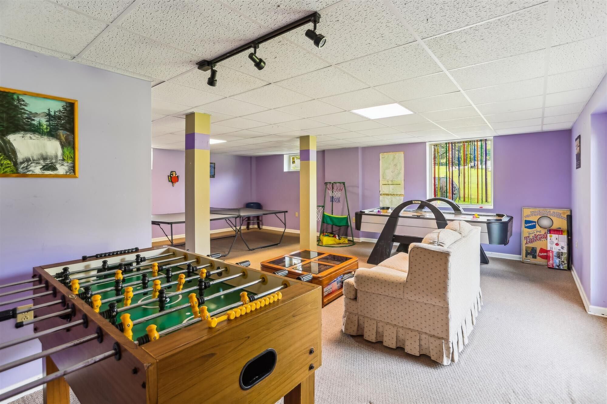 A room with a foosball table, a sofa, a basketball hoop, and a window with a view of trees.