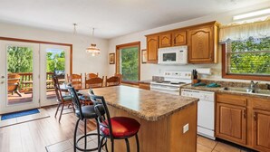 A kitchen with wooden cabinets, a granite countertop, a built-in microwave, a stove, and a sink. There is a dining area with a table and chairs, and a deck with a view of trees. - Enjoy some mountain time with your favorite canine at Alpine View! (Dry Fork)