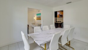 A dining area with a marble table and white chairs, a kitchen in the background, and a view of the living room. - Luxe Wimauma Home w/ Pool & Lake Views! (Wimauma)