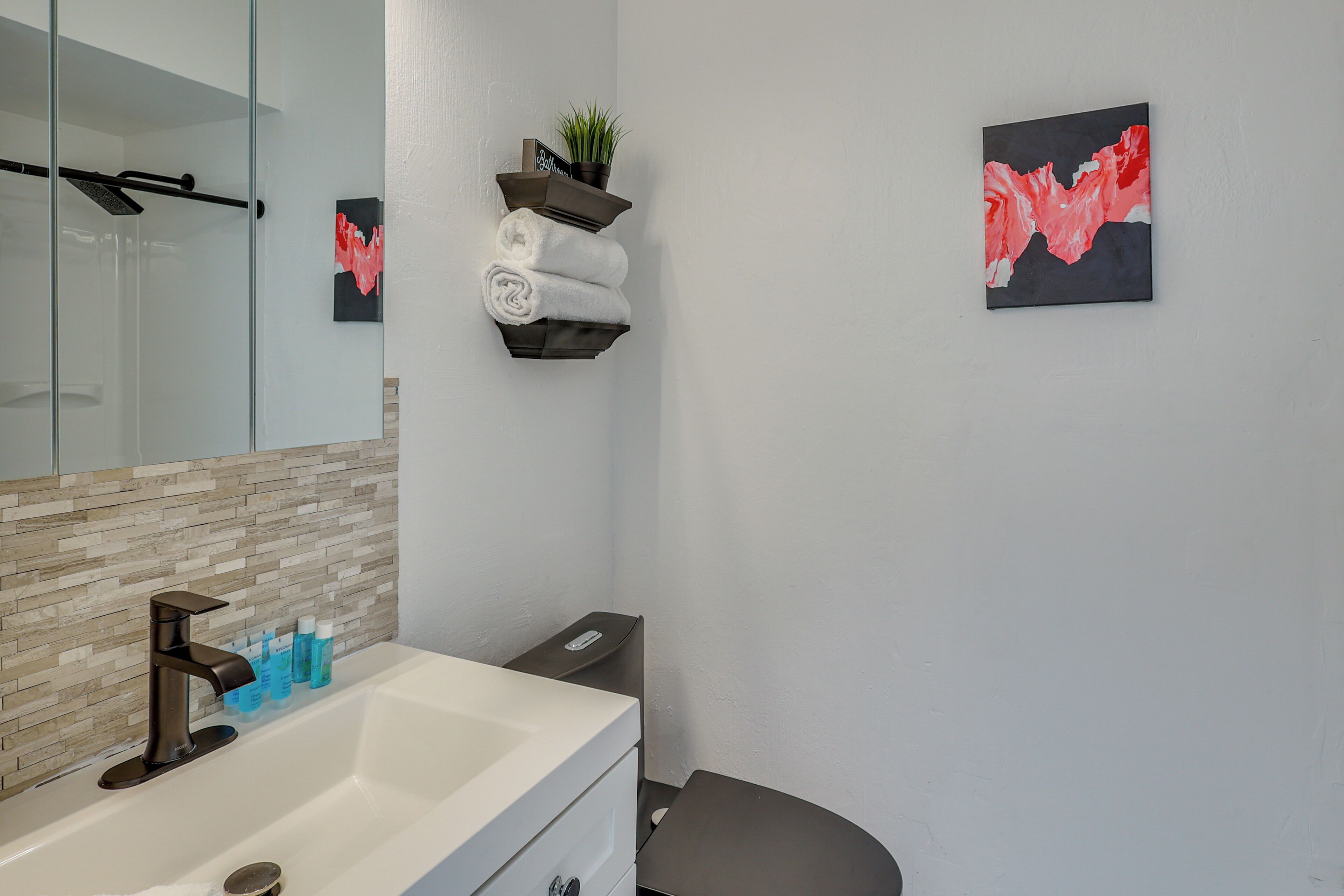 A bathroom with a glass shower enclosure, a white sink with a modern faucet, a towel rack with folded towels, a small potted plant, and two abstract paintings on the wall.
