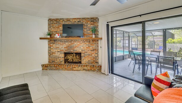 A living room with a brick fireplace, a flat-screen TV, a ceiling fan, and a large sliding glass door leading to an outdoor patio. - Luxe Wimauma Home w/ Pool & Lake Views! (Wimauma)