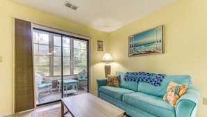 Una sala de estar con un sofá color turquesa, una mesa de centro de madera, una lámpara y un cuadro con una escena de playa.